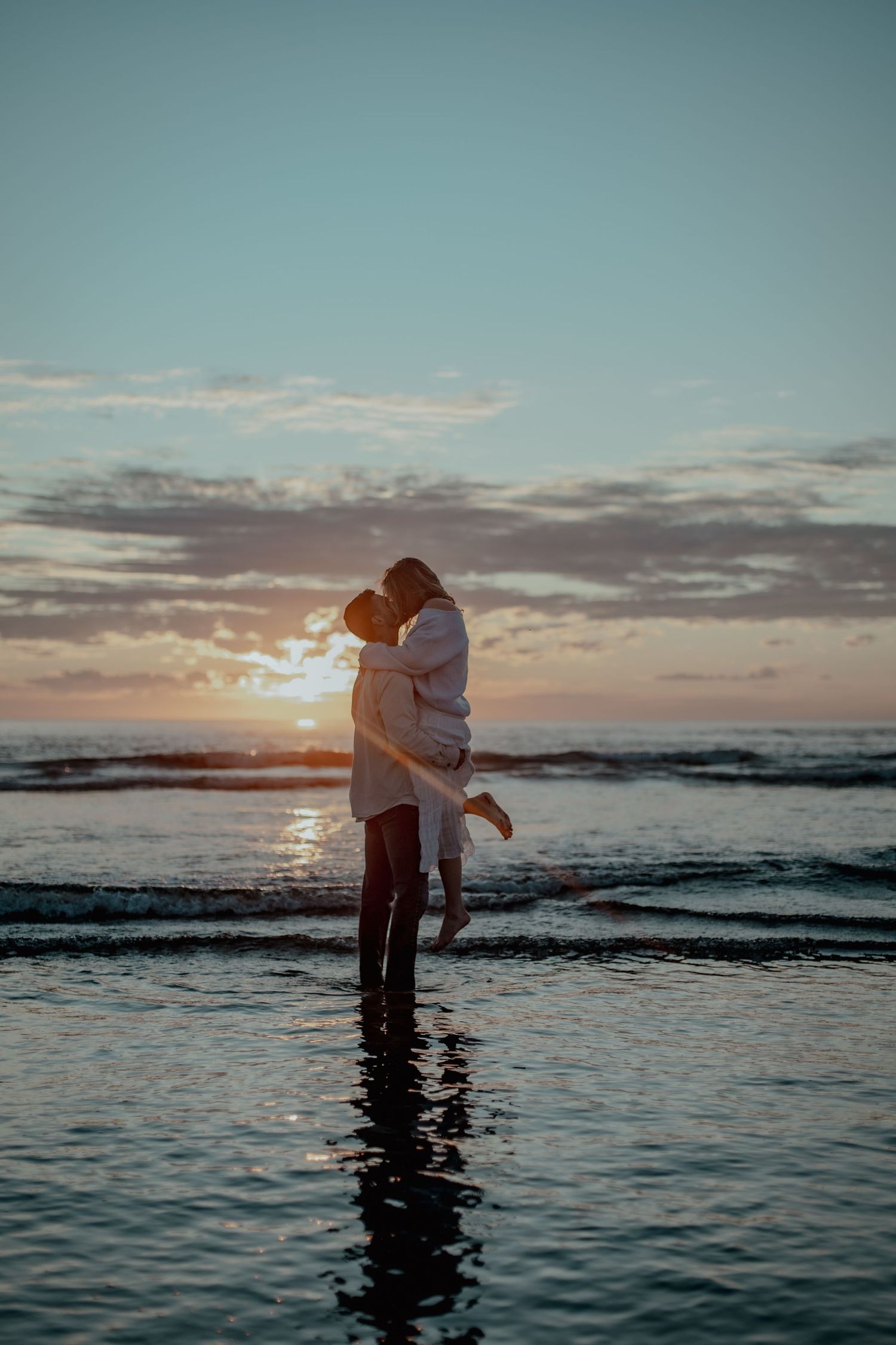 A couple embraces while standing in shallow ocean water during a romantic sunset at the beach.