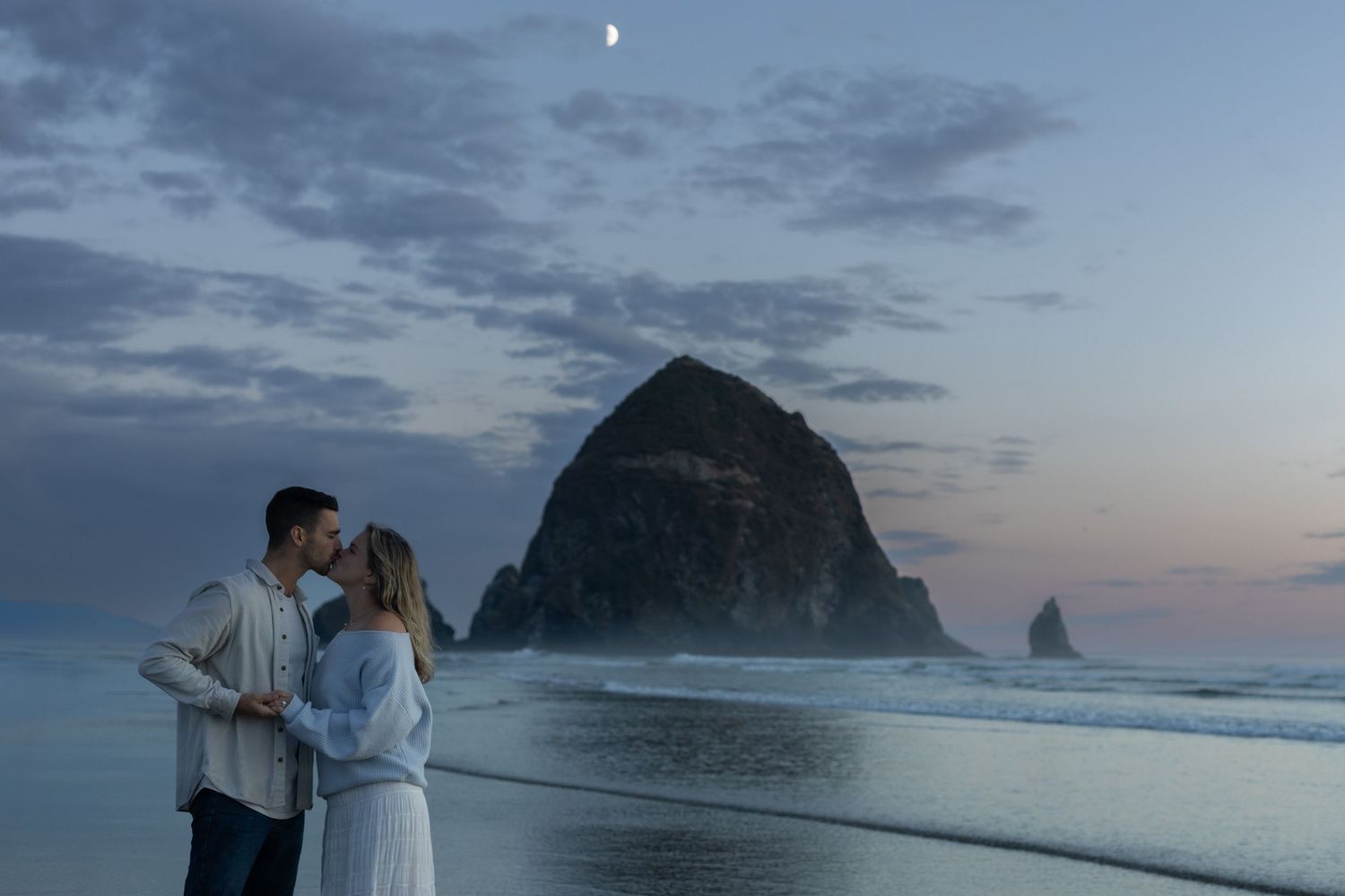 A couple stands together on a misty beach at dusk with Haystack Rock silhouetted against the twilight sky.