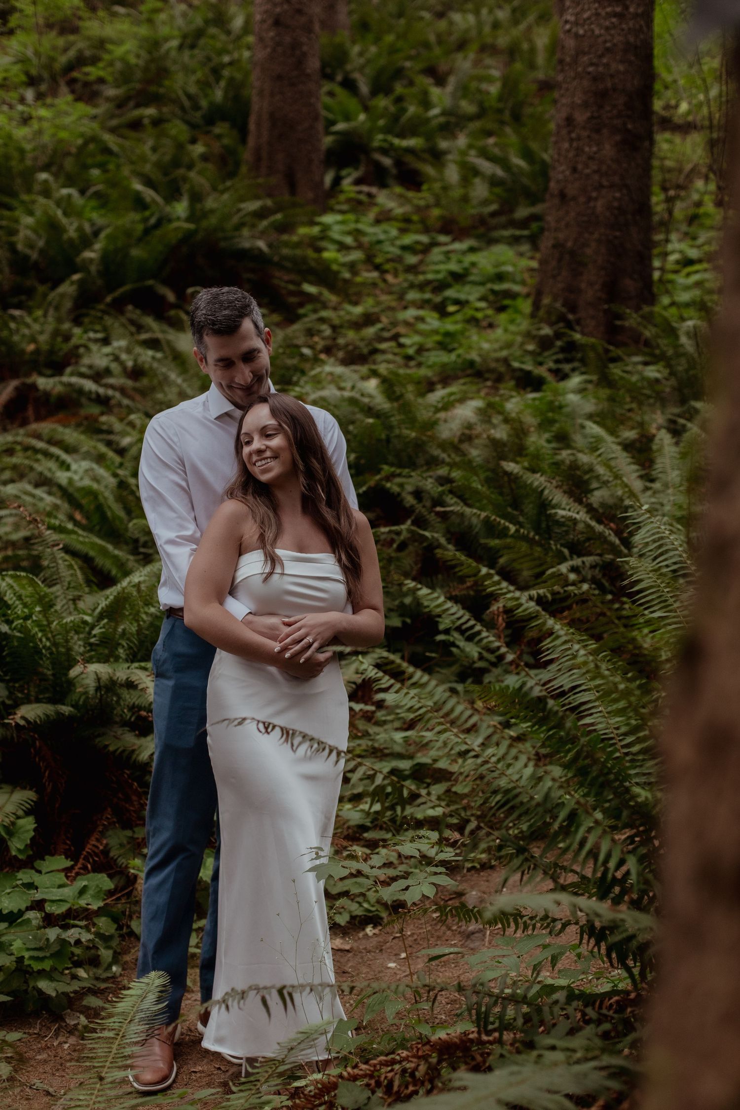 A romantic couple embraces in a lush forest setting surrounded by ferns and tall trees during a wedding photoshoot.