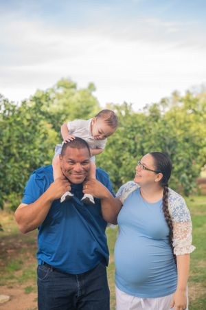 A joyful outdoor family portrait showing parents and a toddler sitting on shoulders with green trees in background.