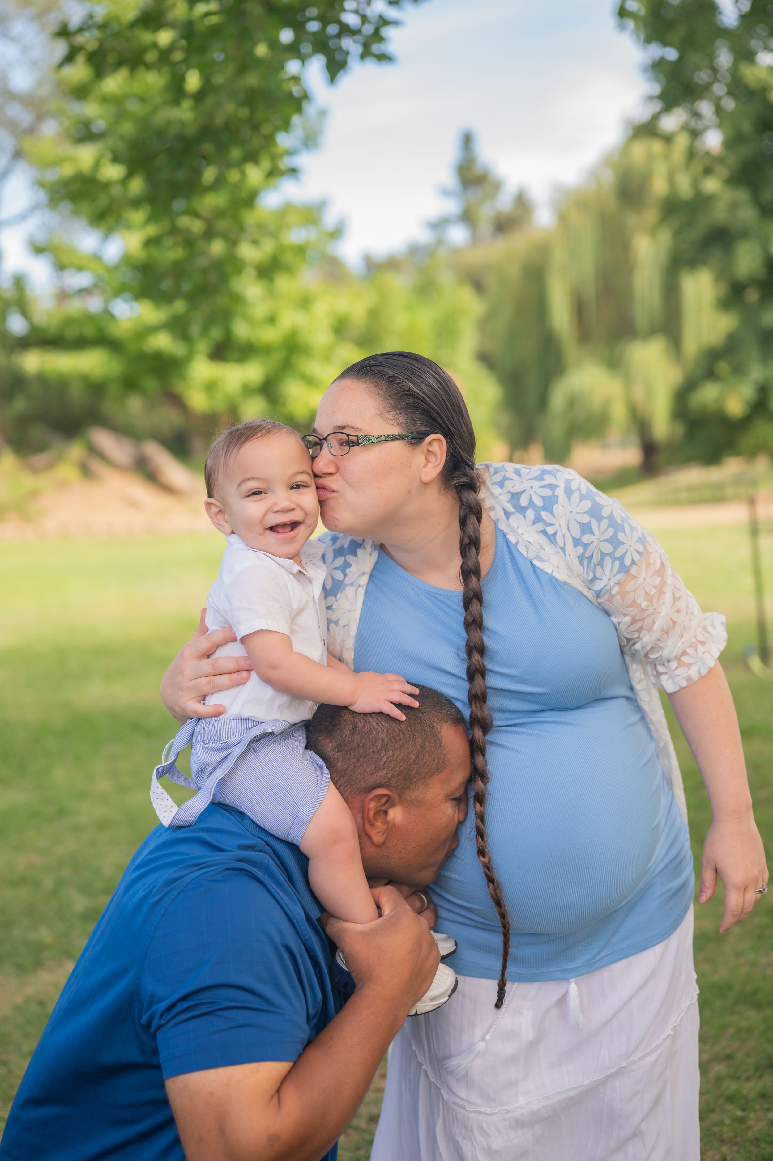 Tender family moment captured outdoors with expecting parents and joyful baby against green landscape.