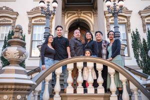 A group of people stand together on ornate stone steps with decorative balustrades and classic lampposts behind them.