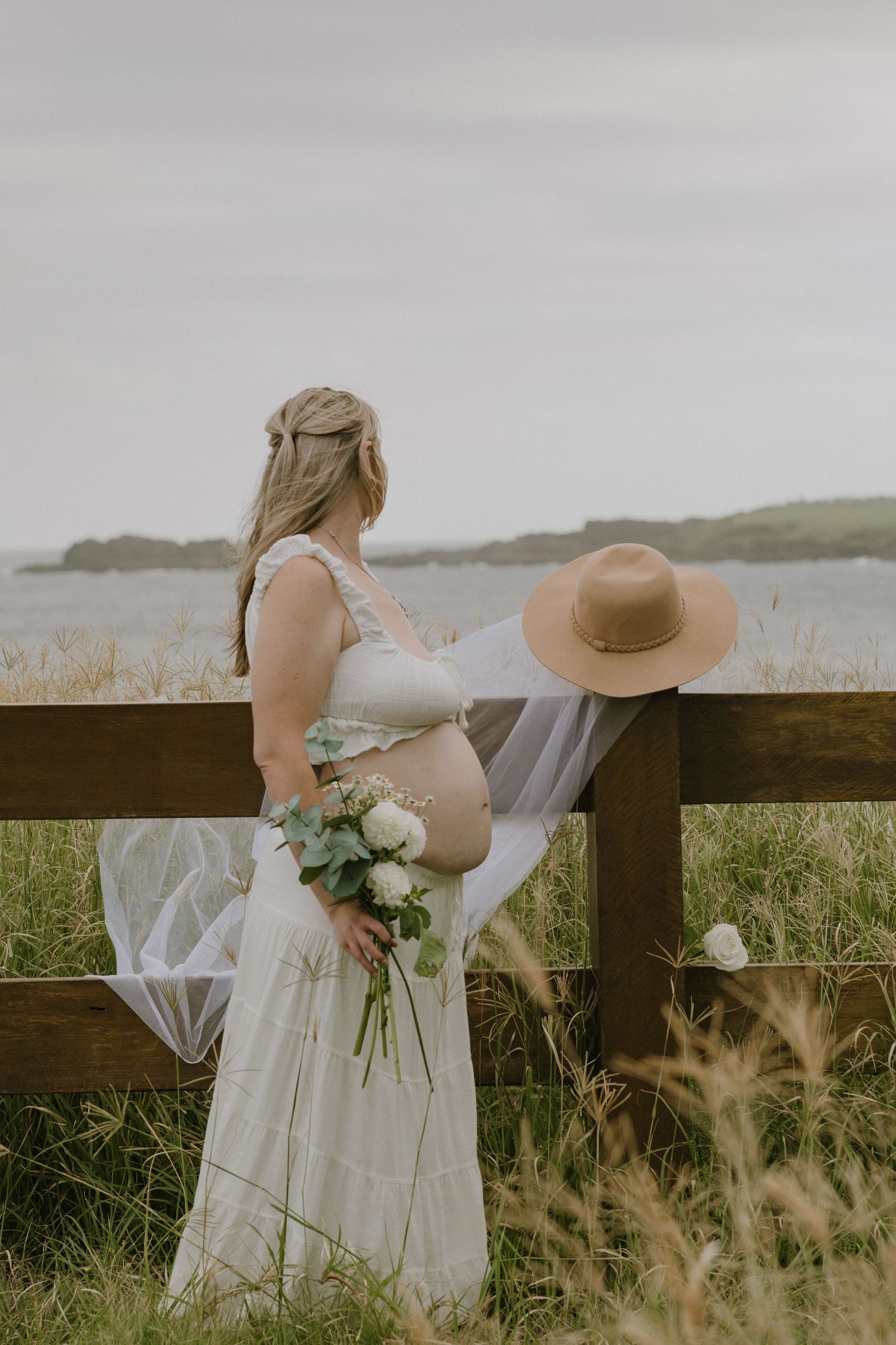A person in a wedding dress holding flowers stands by a wooden fence overlooking water.