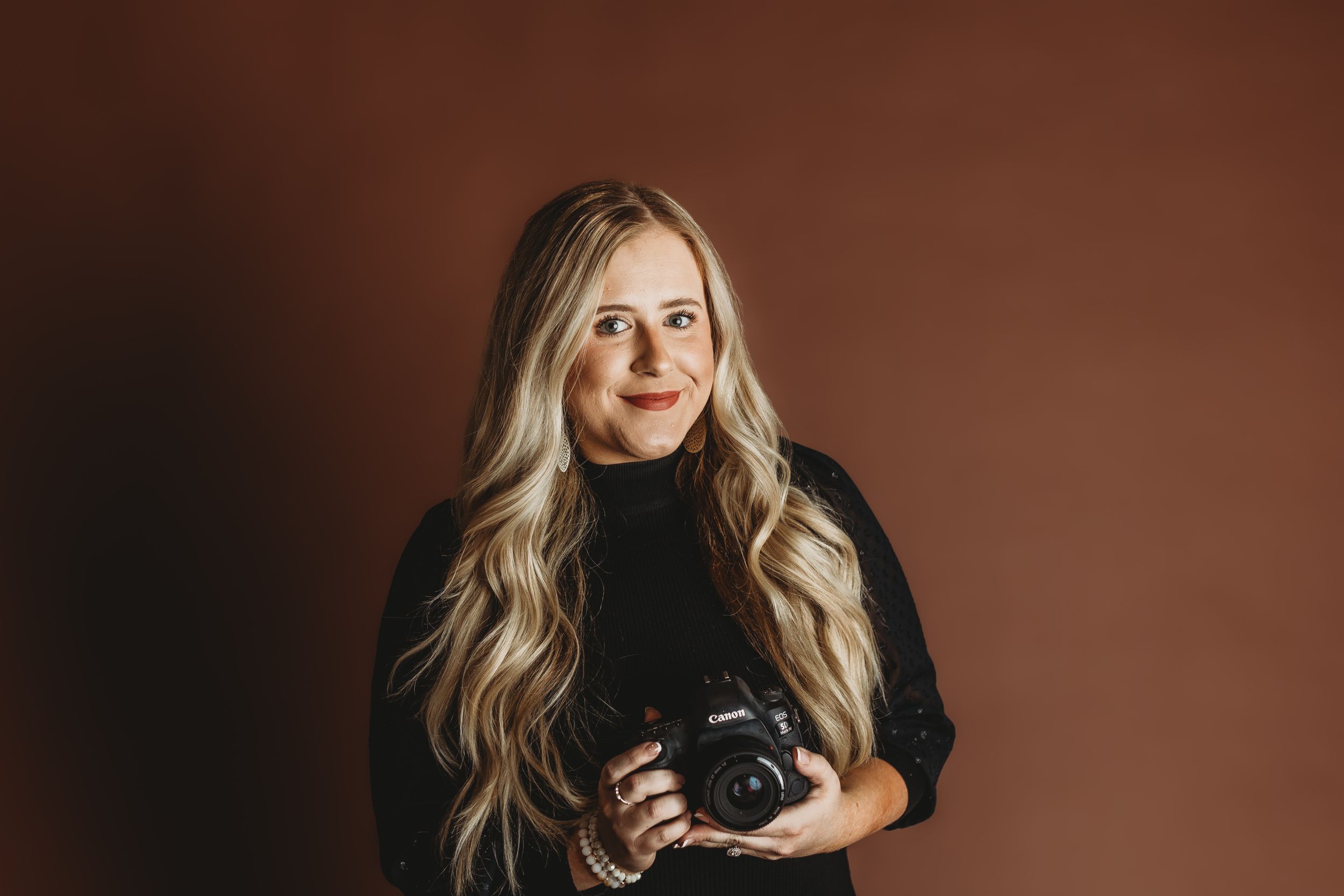 Professional photographer in black sweater holds camera against brown studio background.