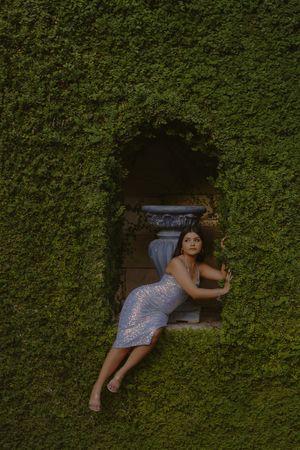Woman in lace dress posing elegantly in an ivy-covered wall niche with a decorative stone urn backdrop.