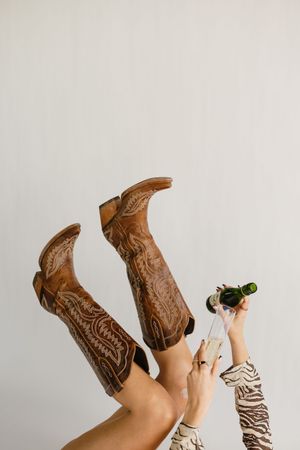 Woman in brown cowboy boots with legs raised, pouring champagne into a glass, wearing a zebra print top.