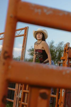 Young woman in white cowboy hat and black top poses among rustic orange metal ranch gates outdoors.