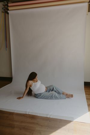 Pregnant woman in white crop top and blue jeans reclining on white backdrop in photography studio.