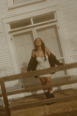 Young woman in white dress and black jacket posing by rustic wooden fence outside vintage white farmhouse.