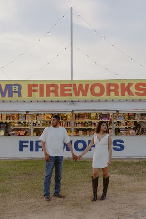 Couple holding hands in front of a colorful fireworks stand, woman in white dress and boots, man in jeans and white tee.