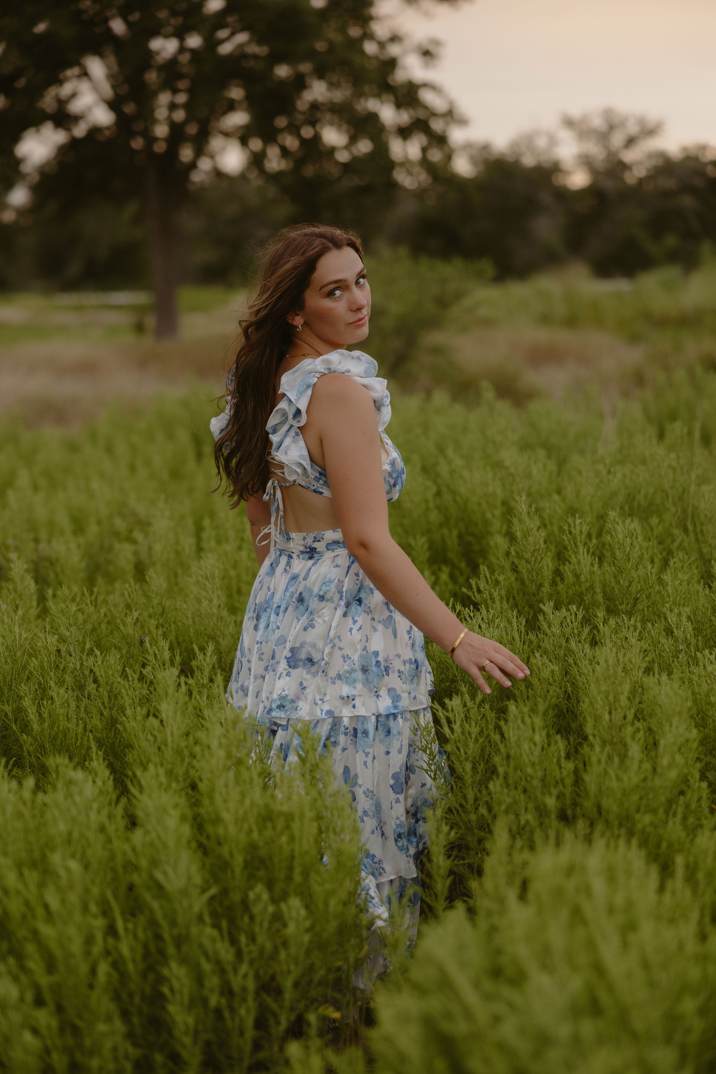 Woman in blue floral dress standing in lush green field at sunset, looking over shoulder.