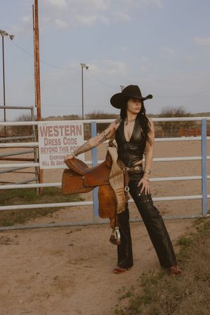 A figure in western attire poses against a rustic ranch backdrop with wooden fencing and a Western dress code sign.