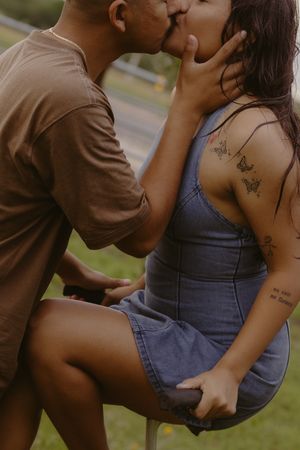 Couple sharing a romantic kiss outdoors, woman in denim dress with butterfly tattoos sitting on a stool.