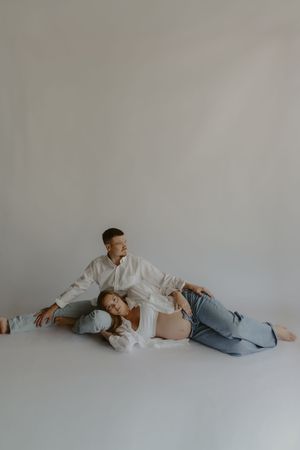 Pregnant couple posing on studio floor, man in white shirt sitting behind woman in jeans showing baby bump.