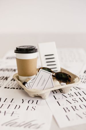 Aesthetic flat lay with takeaway coffee cup, tortoise shell sunglasses, and business card in cardboard tray on lash salon prints.