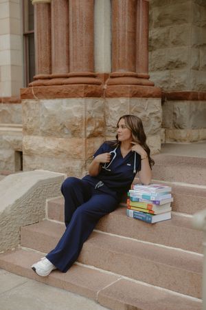 Nursing student in navy scrubs with stethoscope sits on stone steps beside stacked medical textbooks.