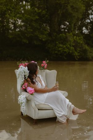 Bride in white dress with floral crown sits in armchair surrounded by floodwater, holding a glass, with green trees behind.