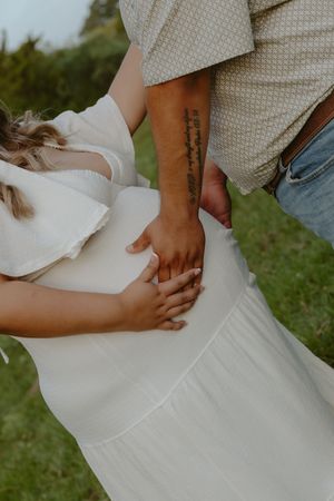 A couple tenderly holds hands over a pregnant belly outdoors, the man's tattooed arm visible against her white dress.