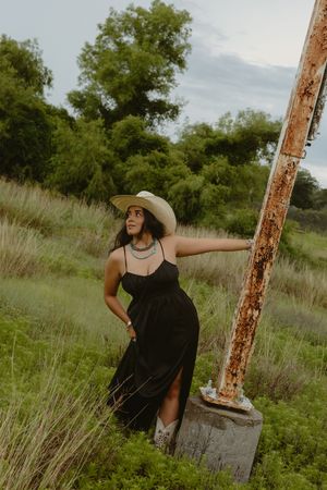 Woman in black dress, wide-brim hat, and boots poses by rusty pole in lush green field.