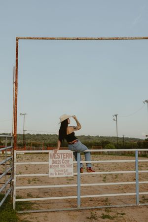 Woman in white cowboy hat and jeans sitting on a ranch gate with a Western Dress Code sign.
