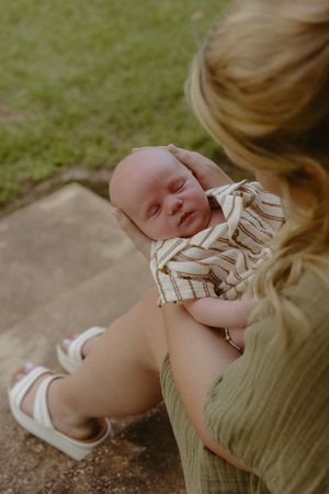 A blonde mother in a green dress gently cradles her sleeping newborn baby outdoors on a sunny day.