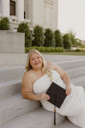 A smiling graduate in a white dress holds her cap on stone steps, celebrating her graduation outdoors.