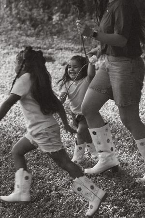 Black and white photo of two girls playing on a rope swing outdoors, with an adult in cowboy boots supervising.