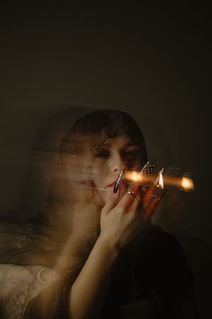 A mysterious woman with dark nails lights candles in a moody, dimly lit setting with a ghostly double exposure effect.
