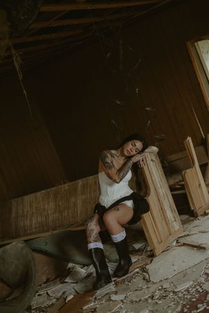 Tattooed woman in white top, thigh-high socks, and black boots sits pensively in a rustic, abandoned wooden interior.