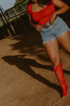 Woman in red bodysuit, denim cutoff shorts, silver belt buckle, and red cowboy boots posing on dirt ground.