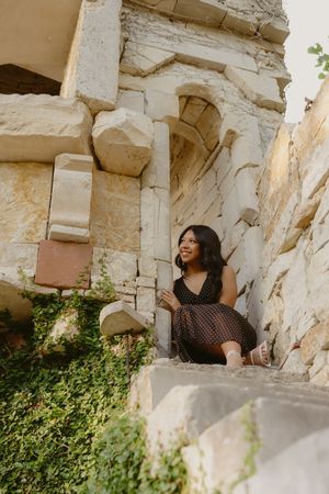 Young woman in dark dress sitting on ancient stone steps, smiling among historic ruins with ivy-covered walls.