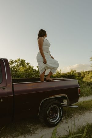 Woman in white dress standing on the bed of a vintage brown pickup truck at sunset with green trees in background.