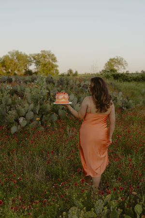 Woman in orange dress holding a cake in a wildflower field with cacti at golden hour.