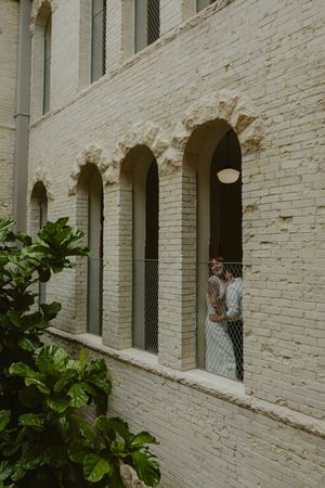 Couple embracing in arched window of cream brick building with lush green foliage in foreground.