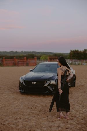 Woman in black dress standing beside a black Cadillac sedan at sunset on sandy ground.
