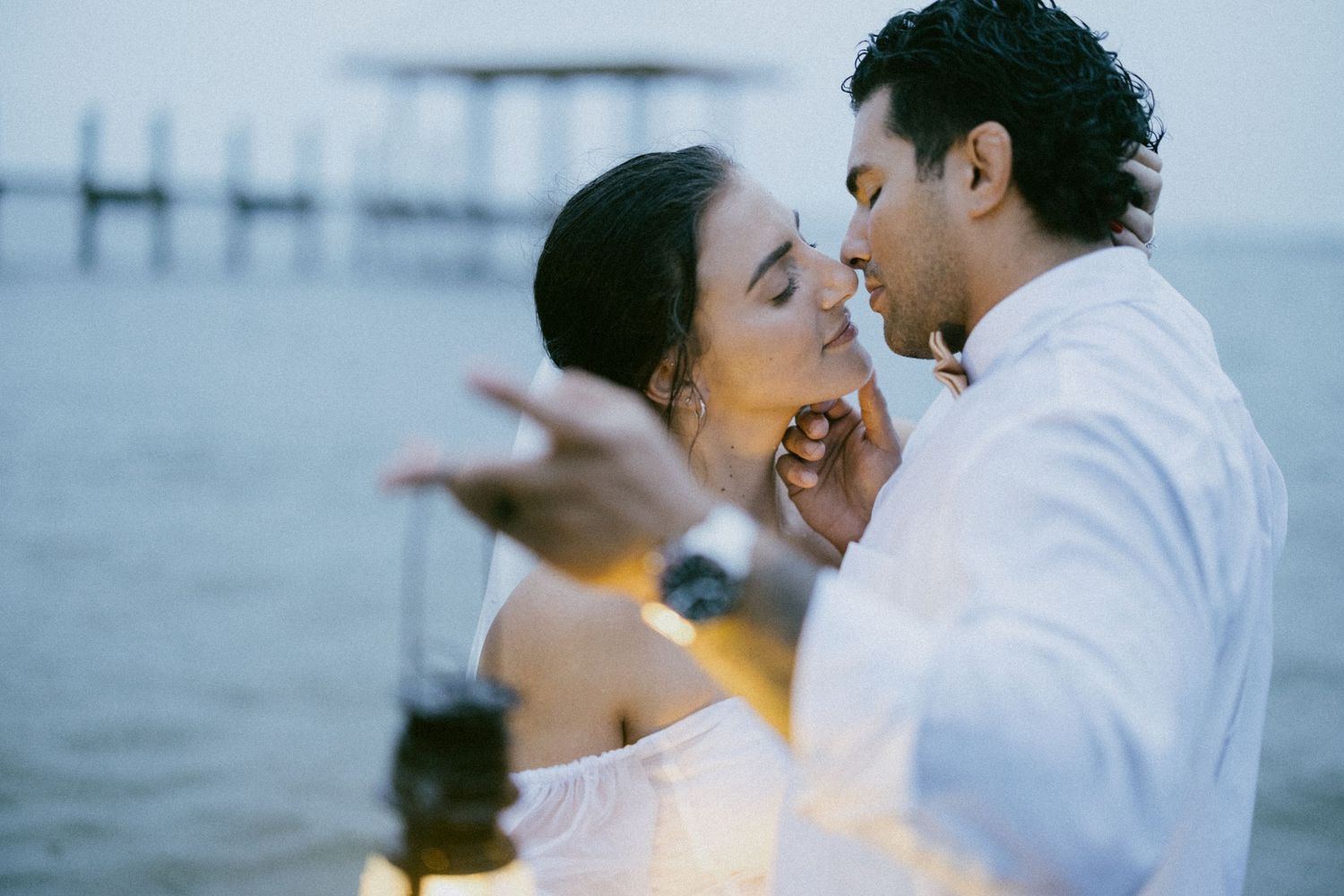 Romantic newlywed couple embracing by pier at dusk with aqua blue ocean waters in background.