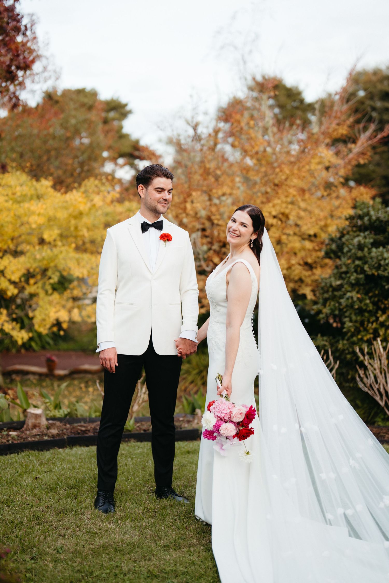 A couple in wedding attire poses against vibrant autumn foliage with pink flowers.