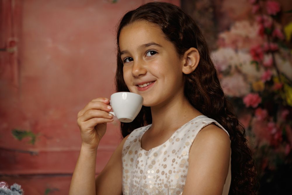 A smiling youth enjoys a warm beverage from a tiny cup against a rosy background with flowers.