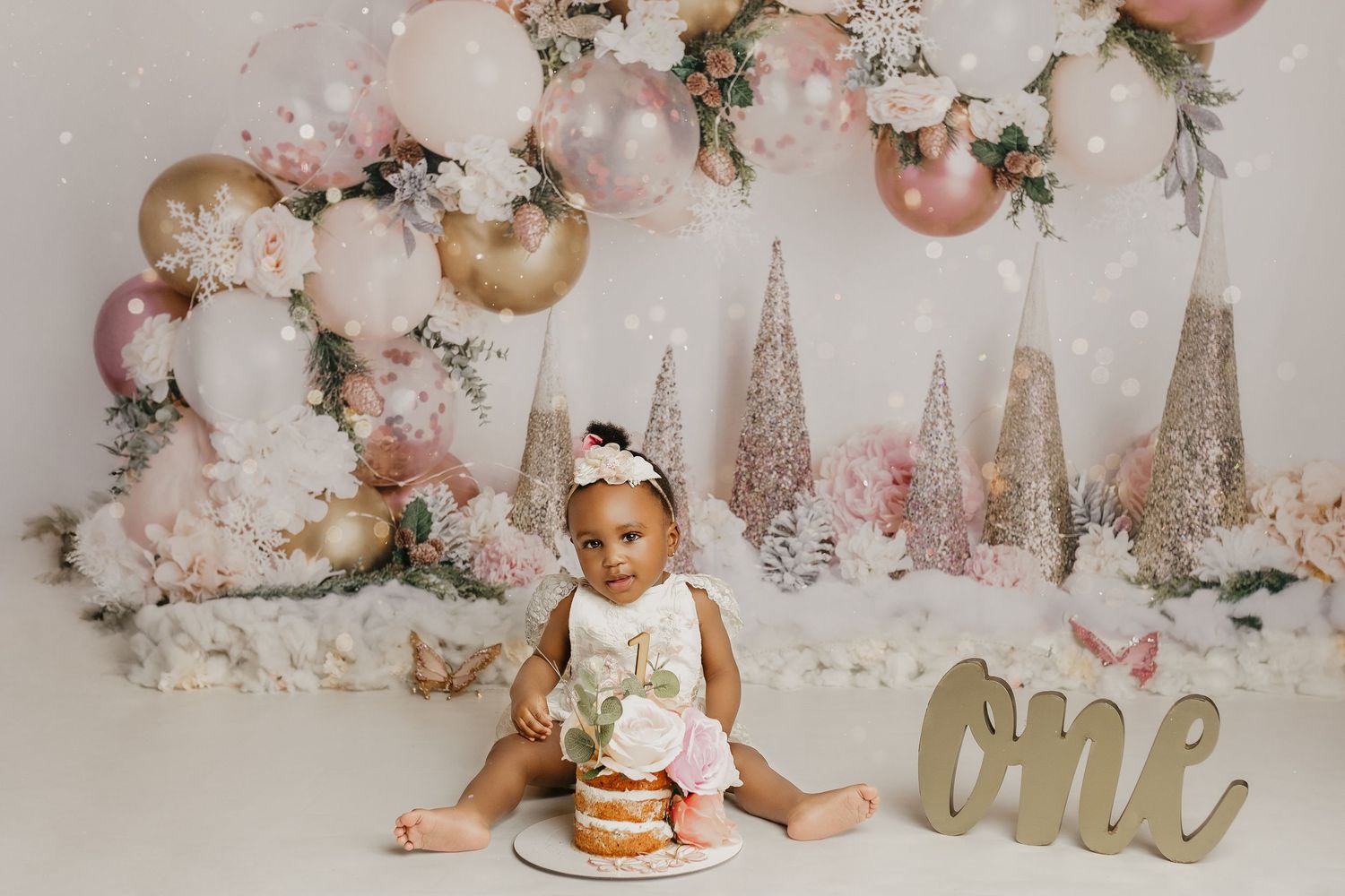 Festive first birthday setup with pink and gold balloons, glittery trees, and a smash cake against white backdrop.