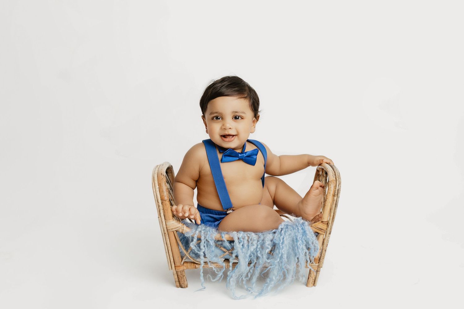 Baby wearing blue outfit sits in wooden basket against white background.