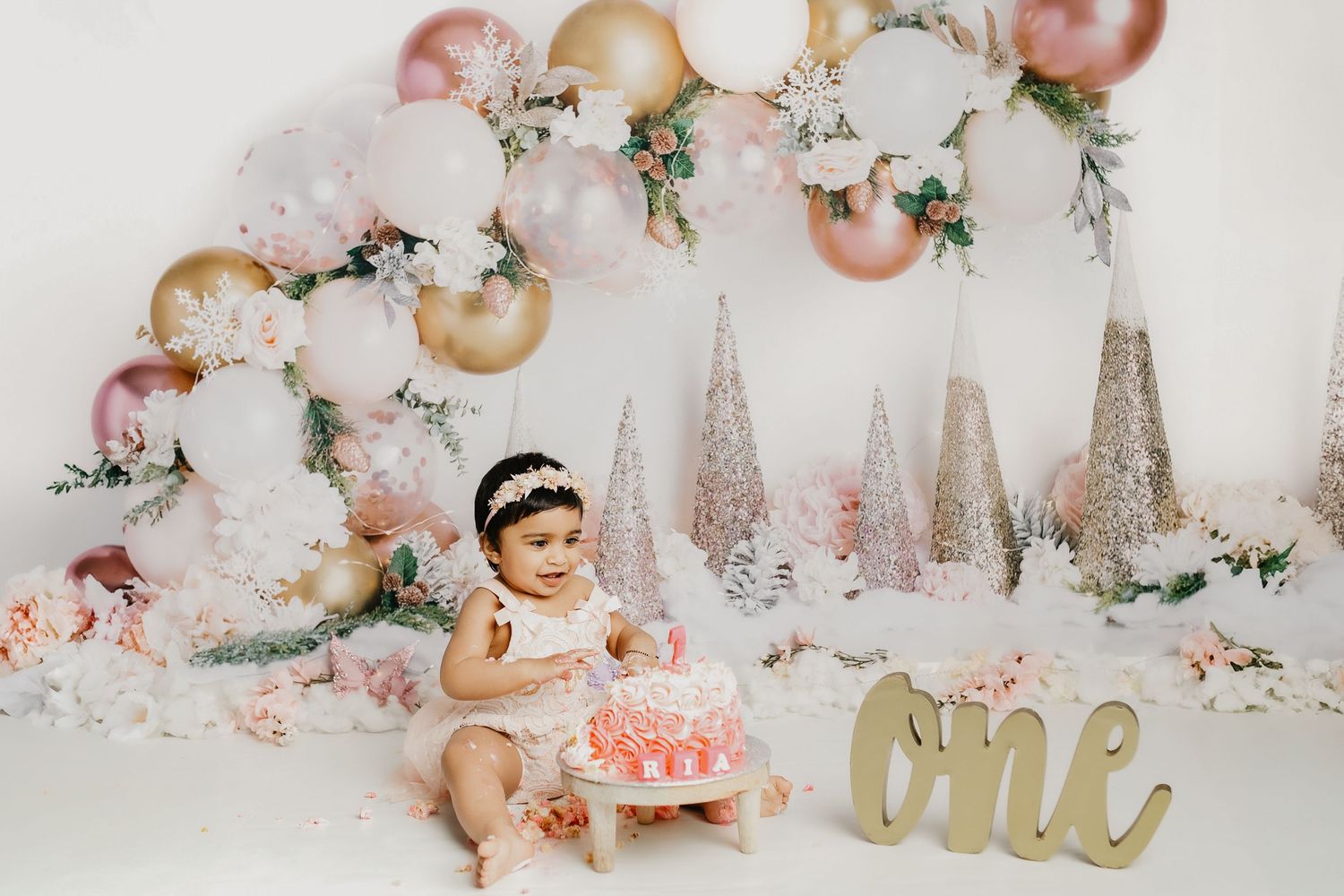 Pink and gold themed cake smash photography setup with floral arrangements and metallic accents.