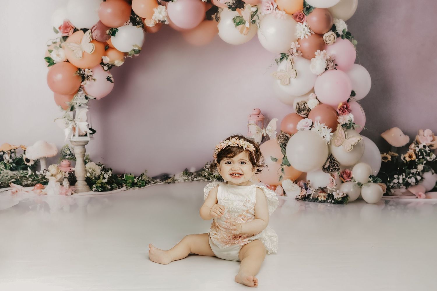 A smiling baby girl in a white outfit sits before a floral balloon arch in pink, white, and peach tones.