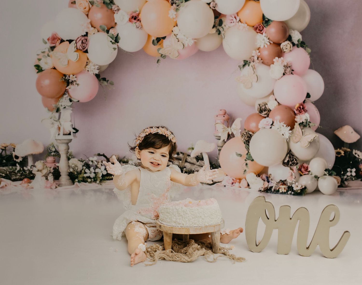 Baby girl in white dress celebrates first birthday with cake smash, surrounded by pink and white balloon arch.