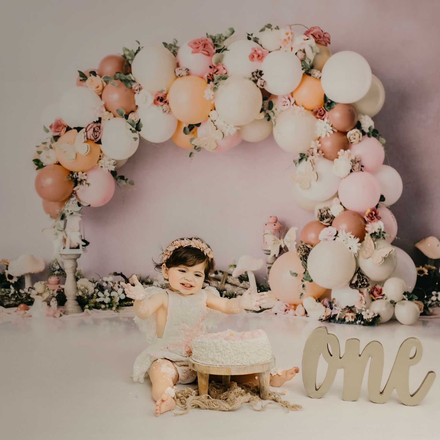 First birthday cake smash setup with pink and white balloon arch and floral decorations.
