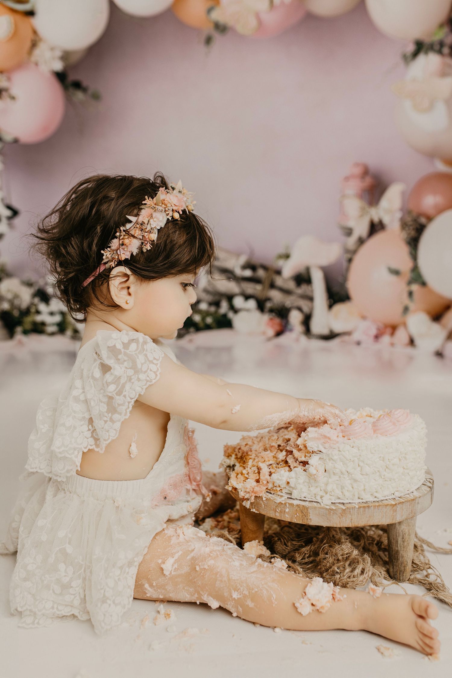 A baby girl in a white lace dress smashes her first birthday cake surrounded by pink balloons and floral decorations.