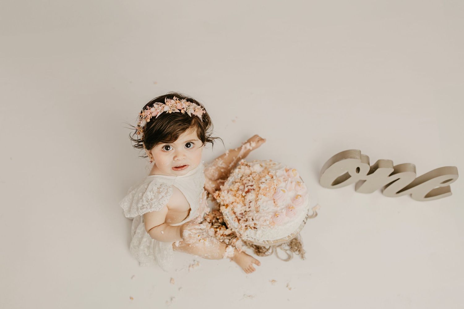 Baby girl in white dress and floral crown sits with a pink peony cake smash, next to a wooden 'one' sign.