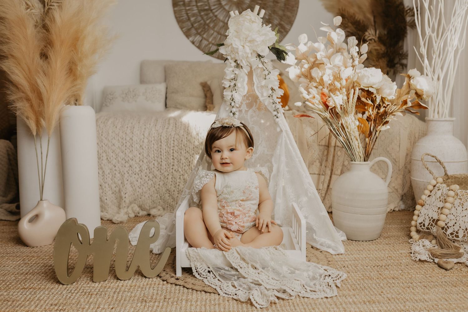 Baby girl in floral romper sits by boho teepee with pampas grass and white florals for first birthday photo.