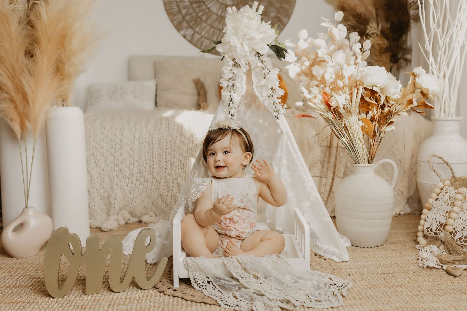 Baby girl in boho floral first birthday photo shoot sitting in mini teepee surrounded by pampas grass and white vases.
