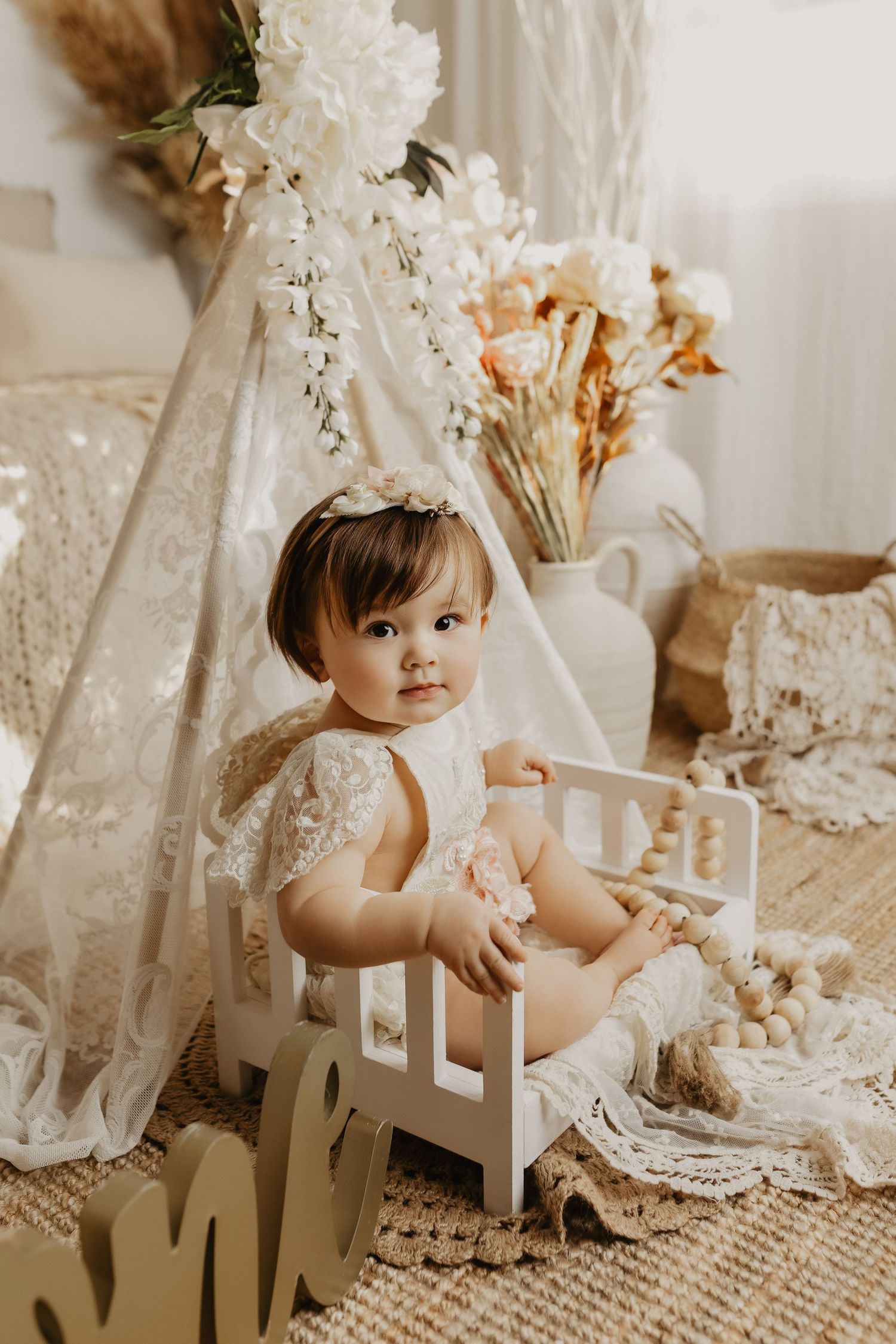 Baby girl in lace romper sits in white crib with boho floral teepee backdrop.