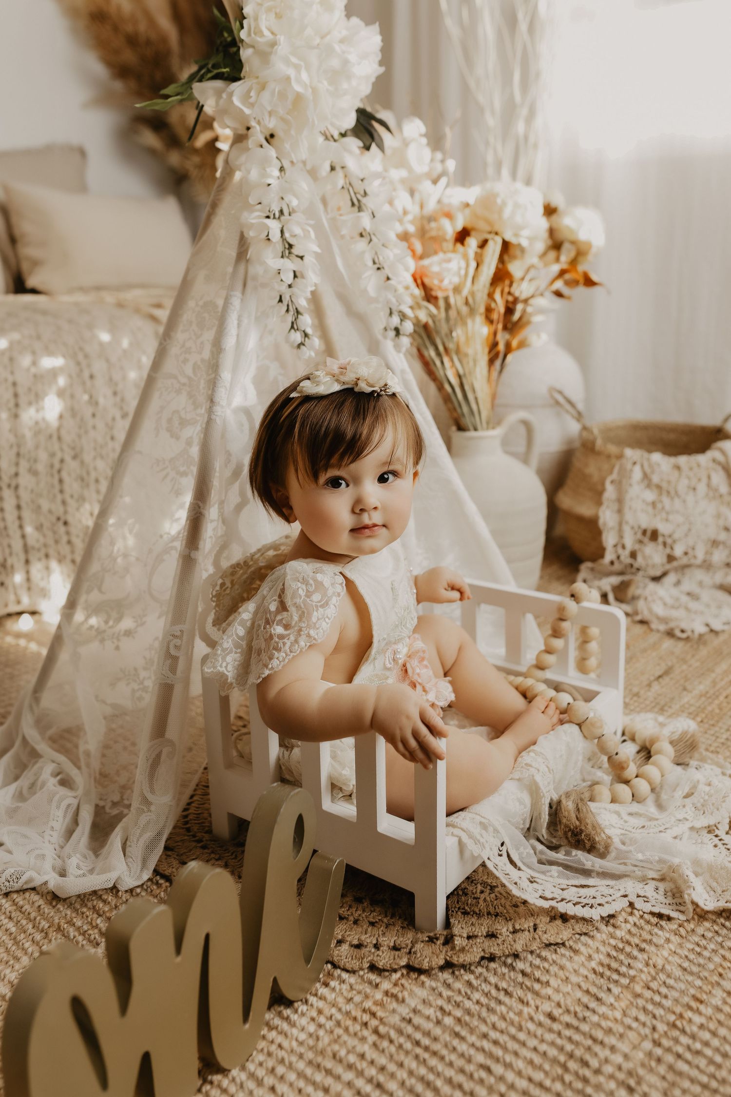Baby girl in lace dress sits in boho first birthday setup with teepee, flowers, and wooden ONE sign.